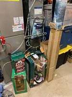 Various boxed Christmas holiday decor items arranged near basement utilities including hobby Santa figure, potpourri pot Santa workshop figure, boxed snowman light silhouette, and boxed staked Christmas balls.