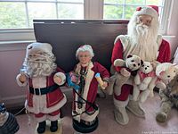 Photo showing three Christmas figures: two Santa Claus and one elf-like figure posed in front of a window. Tall Santa holds plush teddy bears. The figures have fabric, plastic features, and some missing light bulbs.