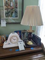 Photo showing a wood and brass desk lamp with pleated cream fabric shade beside a white ceramic mantle clock with floral design and gold-tone bezel.