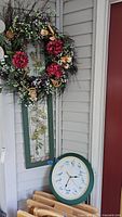 Wood framed vertical stained glass window panel with two roses design and colorful wreath of artificial flowers and berries hanging on a wall next to a clock.