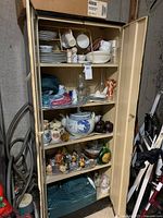 Metal shelving unit with five shelves holding assorted collectibles including figurines, plates, cups, glassware, and a large teapot urn visible in center shelves.