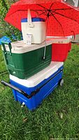 Stack of five coolers including a large blue cooler on the bottom, green cooler, small white cooler with blue lid, a red cooler, and white cooler on top with a red umbrella providing shade.
