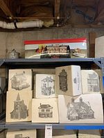 Photo showing eight Dickens Village house boxes with detailed black and white illustrations stacked on a metal shelf, with visible packaging and some wear.