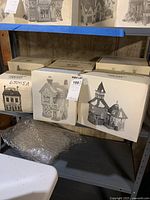 Six cardboard boxes each showing black-and-white photographic images of different Dickens Village ceramic houses with snow accents, arranged on a shelf with bubble wrap below them.