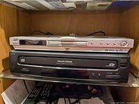 Silver JVC DVD player placed on top of black Philips Magnavox CD changer inside a wooden shelf
