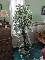 Full view of artificial plant with white flowers in woven basket next to window and furniture.