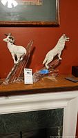 Photo showing both big horn sheep and wolf figurines on angular clear glass bases placed on a wooden surface against red wall.