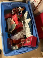 Top view of blue plastic bin containing assorted holiday decorations including Clothtique Santa figurines, a snow-covered decorative item, and red Waterford ornament boxes.