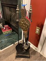 Five wrought iron fireplace tools in holder with decorative brass fire screen element, shown near fireplace and wall outlet.