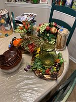 Wide view of all items on table including artificial fruit and floral arrangements, ceramic hen on nest, ceramic bird planter, green glass vase on floral wreath and metal tray.
