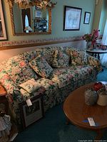 Front view of floral sofa with three cushions and four matching pillows, floral arm covers draped on armrests. Sofa situated in a living room setting with paintings on wall and wooden coffee table in front.