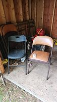 Four metal folding chairs with padded vinyl seats and backs (2 brown, 2 black) in a wooden shed on a concrete floor with tools and miscellaneous items around.