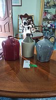 Front view of three ceramic jugs on wooden table, different colors: burgundy, beige with green glaze, gray. Background includes chair and cabinet with collectibles.