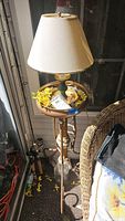 Full view of plant stand holding a table lamp with white shade and some yellow decorative flowers inside the lamp base area.