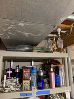Two shelves with assorted items including lamps, dishware, tins, water bottles, and containers arranged in no particular order.