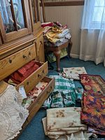 Photo showing open wooden drawers and linens spread on chair and floor with folded cloths of various colors and patterns.