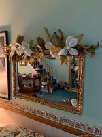 Full view of the ornate gold framed mirror with white floral arrangement on top, showing its position above a couch and floral wallpaper border.