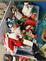Photo of plastic bin containing assorted Christmas plush figurines including snowmen, penguins, elves, and Santa hat