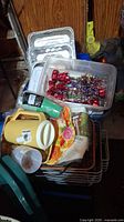 Photo showing assorted disposable aluminum trays, various baskets, a yellow and white cup, and party decoration items such as foil gift bows in plastic bin.