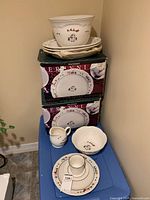 Dinnerware set with original boxes stacked, showing large serving bowl, oval platters, smaller bowl, creamer jug and plates arranged on a table