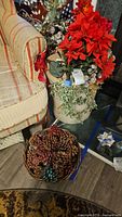 Wicker basket filled with assorted pine cones including some colored red and turquoise, placed next to ceramic planter with artificial Christmas flowers and green foliage around.