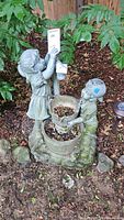 Full view of plastic outdoor fountain depicting two child figures holding water buckets, placed on the ground outdoors surrounded by greenery.