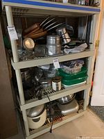 Three shelves of mixed kitchenware showing pans, grinders, containers, and utensils in plastic shelving unit.