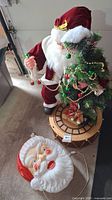 Full view of blow mold Santa on drum base holding Christmas tree and bead garland, with small train on drum. Separate large blow mold Santa face with electrical cord on floor.