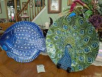 Both serving platters side by side on a wooden table, showing the blue fish-shaped platter on the left and the round green peacock platter on the right.