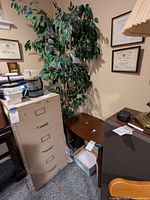 Wooden side table, large faux tree behind it, and part of a filing cabinet visible