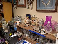 Overview of multiple German steins on shelving, glassware, toby look pitcher, and various decorative items