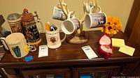 Photo showing three German beer steins, four mugs hanging on decorative faux reindeer antler display, ceramic mug with pens, prayer box and yellow flower arrangement on wooden cabinet
