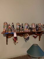 Wide shot of wooden wall shelf showing Norman Rockwell beer steins, Disney figurines, Jim Shore Santas, decorative collector bottles, cat figurines along with hanging dolls and bird beneath the shelf.