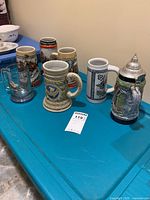 Photo showing six assorted beer steins on a blue surface against a wall, including one with metal lid and one clear glass stein with metal handle.