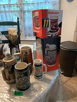Overview of six beer steins, two boxed, placed on a kitchen table with background elements including a window and household items.