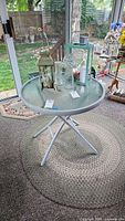 Round table with metal and plastic frame and clear glass top, shown with decorative items placed on top, surrounded by wicker furniture on a patio.