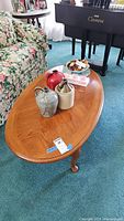 Oval wooden coffee table with medium wood stain. Table surface shown with various decorative items (not included). Table positioned on blue carpet near floral patterned couch and black piano.