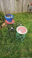 Photo showing two metal framed round tables with colorful mosaic tops, two black metal round planter stands without tops, and one blue ceramic planter pot placed on grass outdoors against a wooden fence.