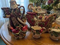 Photo of entire lot on wooden table showing all items included: Fitz and Floyd Santa teapot and cookie jar, cardinal salt and pepper shakers, Cracker Barrel cream and sugar set.