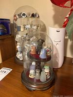 Two display cases with glass domes containing a collection of thimbles, some decorated with floral and emblem designs.
