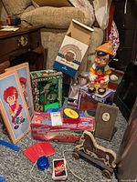 Photo showing collection of various vintage toys and collectibles on carpeted floor by couch, including boxed car, Pinocchio bank, figurines, cards, and wall hangings.