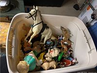 White plastic tub containing assorted toy figurines including people, animals, cartoon characters, and horses.
