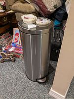 Front view of cylindrical stainless steel trash can with foot pedal, showing two Febreze Scent Stories containers placed on top and some background household items.