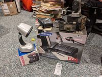 Photo showing Samsung FX40 home fax machine box, Polaroid OneStep Flash camera desk lamp on its box, and a vintage pencil sharpener on carpet with books in background.