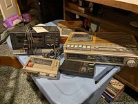 Five vintage radios and cassette players on a blue table including General Electric cassette clock radio, Entroplex traveler FM clock radio, Optimus cassette tape deck, Emerson transistor radio, and Panasonic mini cassette recorder. Cords and antennas visible, items show age.