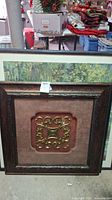 Photo showing the framed decorative metal relief with brown background and ornate frame.