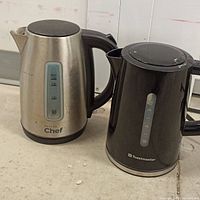 Two electric kettles side by side on a countertop; one stainless steel Master Chef and one black Toastmaster kettle with visible water level indicators