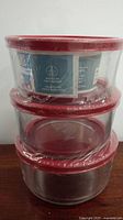 Stack of three round glass containers with red lids, sealed in factory plastic wrap, showing brand sticker affixed