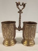 Front view of antique brass double cup showing both cups and decorative handle with etched figures and motifs.