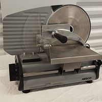 Side view of the Nesco FS-250 food slicer showing the round stainless steel blade, handle, and sliding meat carriage on a gray metal body.